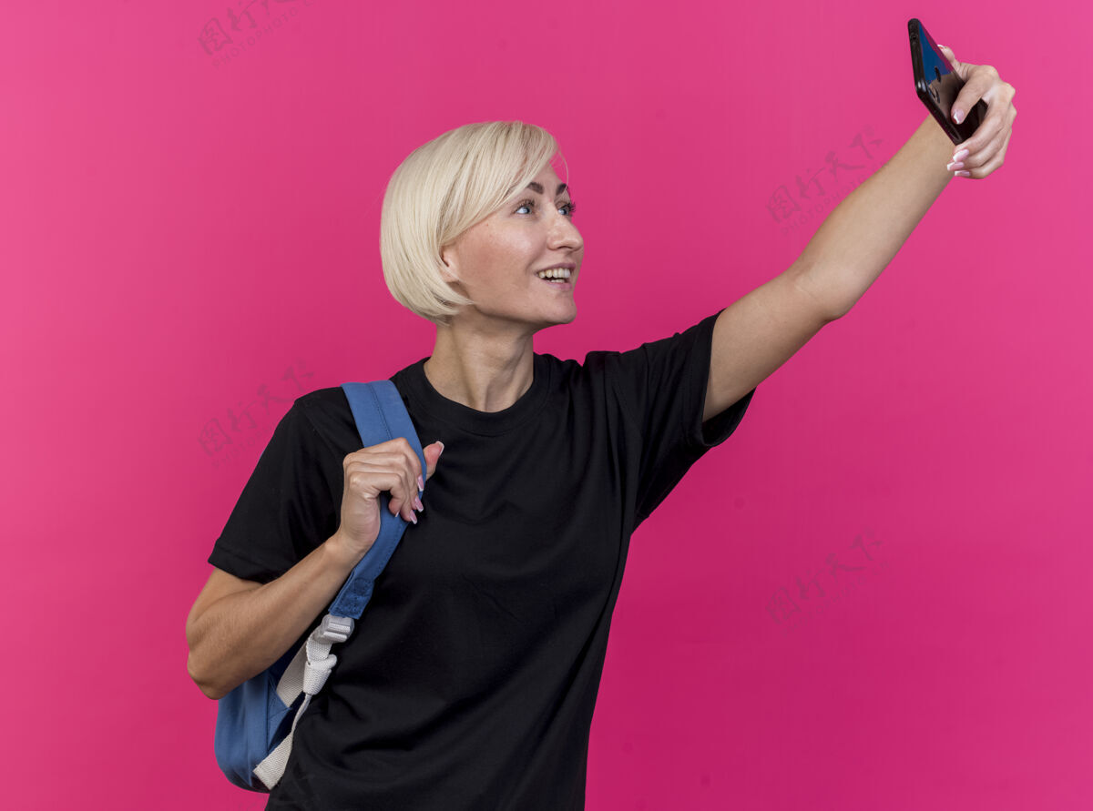 感觉快乐的中年金发斯拉夫女人背着背包拿着背带在粉红色的墙上自拍背姿势金发