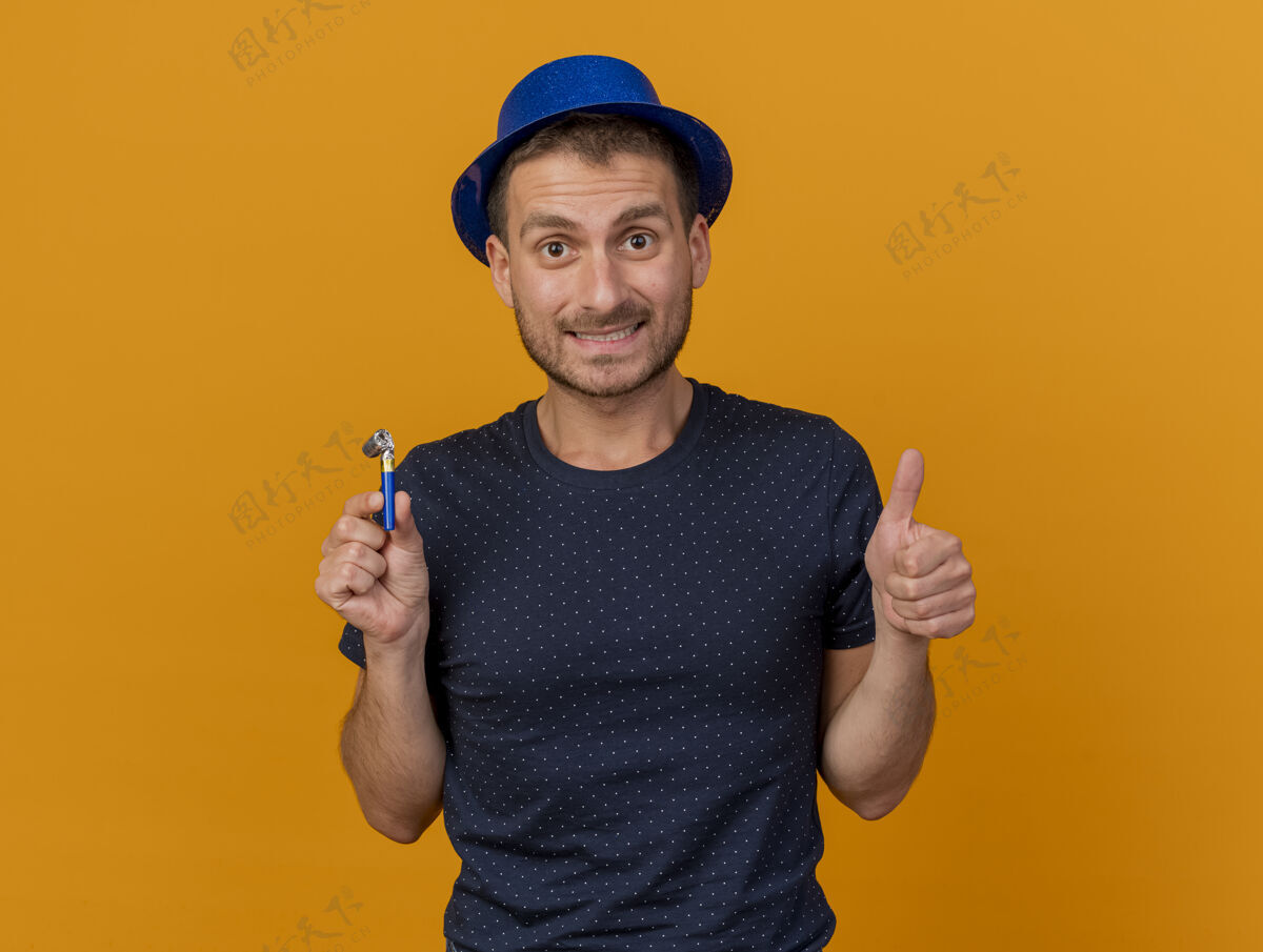 人穿着蓝色派对帽的帅哥竖起大拇指 把派对哨子孤立地举在橙色的墙上 留着复印空间人人英俊
