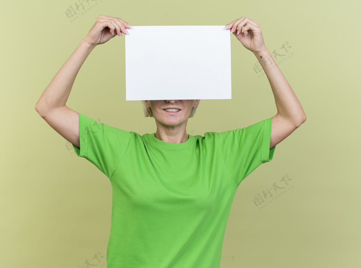 橄榄微笑的中年金发斯拉夫妇女拿着空白纸在眼前孤立在橄榄绿的墙上空白感觉人
