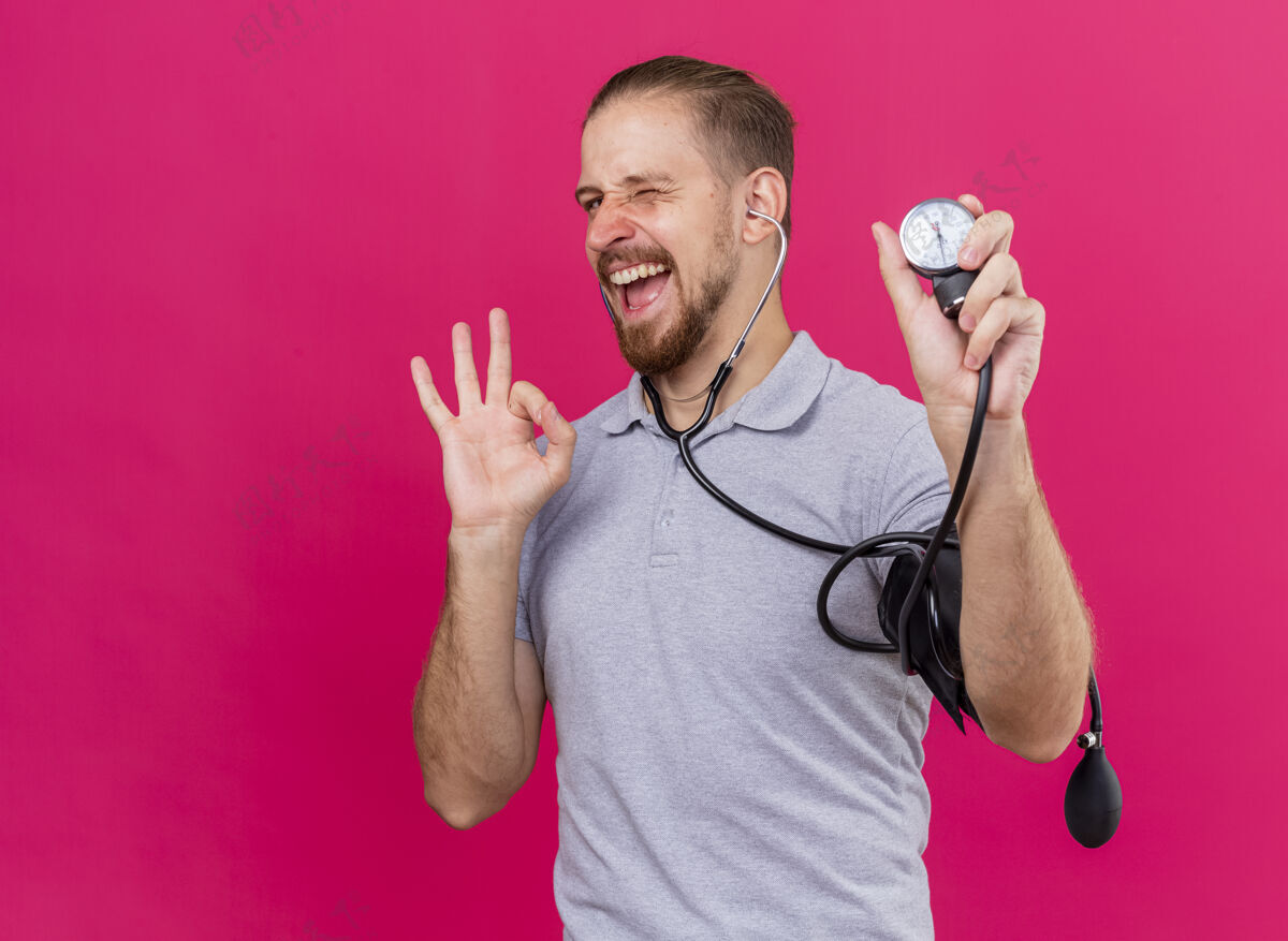 感觉自信的年轻英俊的斯拉夫病人戴着听诊器 用血压计测量他的血压 在前面眨眨眼 做得很好 在粉红色的墙上唱歌 有复制空间人血压计站姿