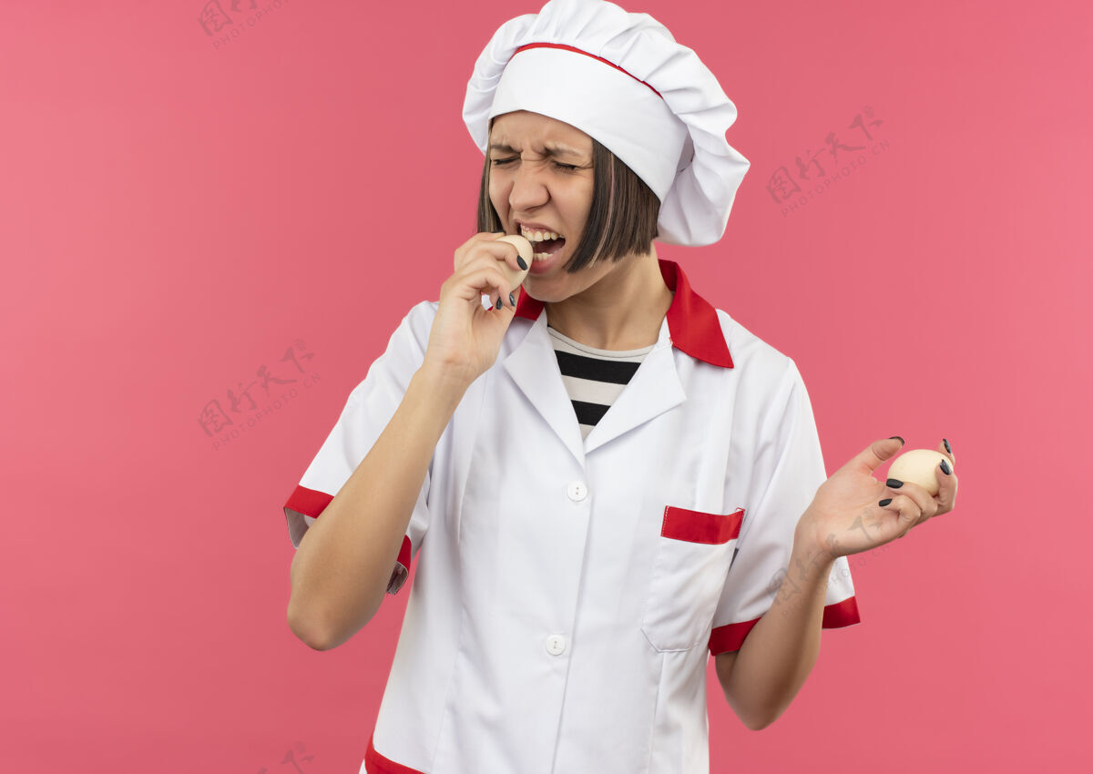 试试年轻的女厨师穿着厨师制服 拿着和试图咬鸡蛋隔离在粉红色背景与复制空间鸡蛋女咬