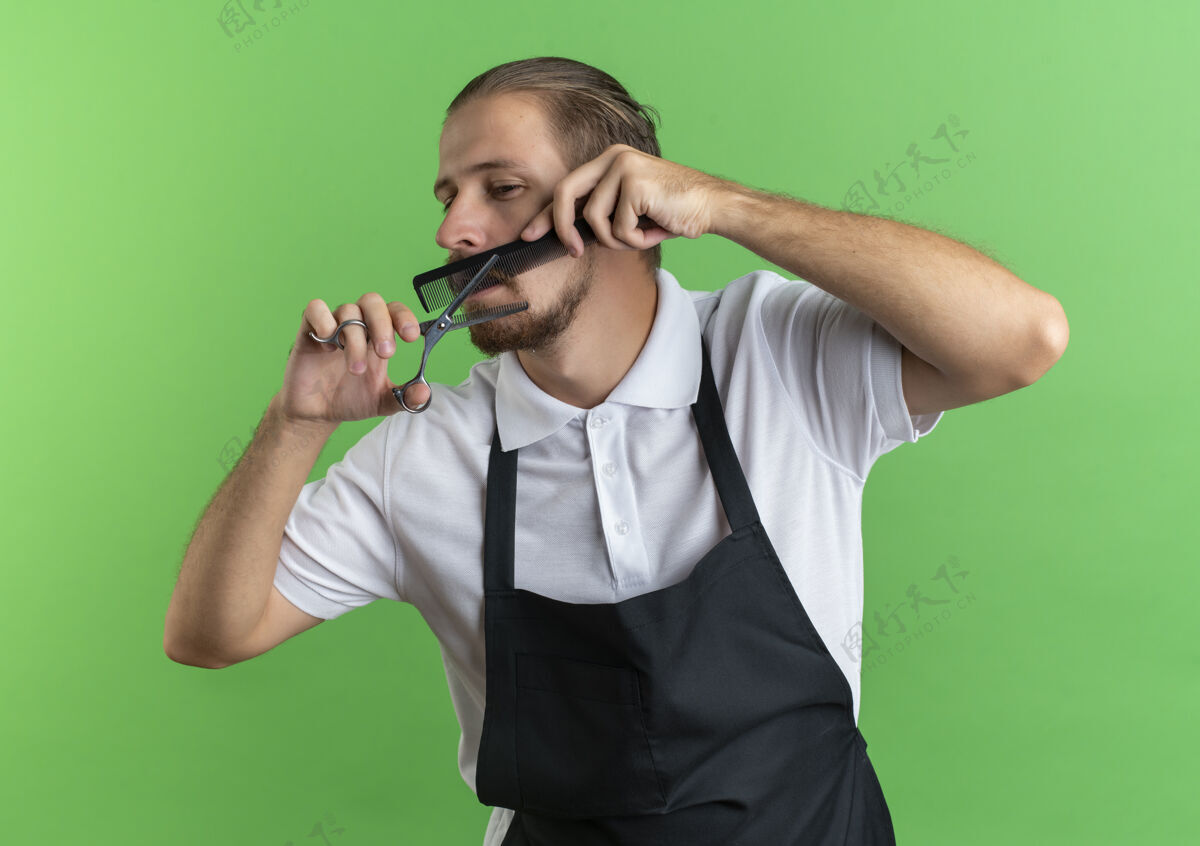 穿年轻英俊的理发师穿着制服 梳着胡子 看着绿色背景上孤立的一面帅气小胡子梳
