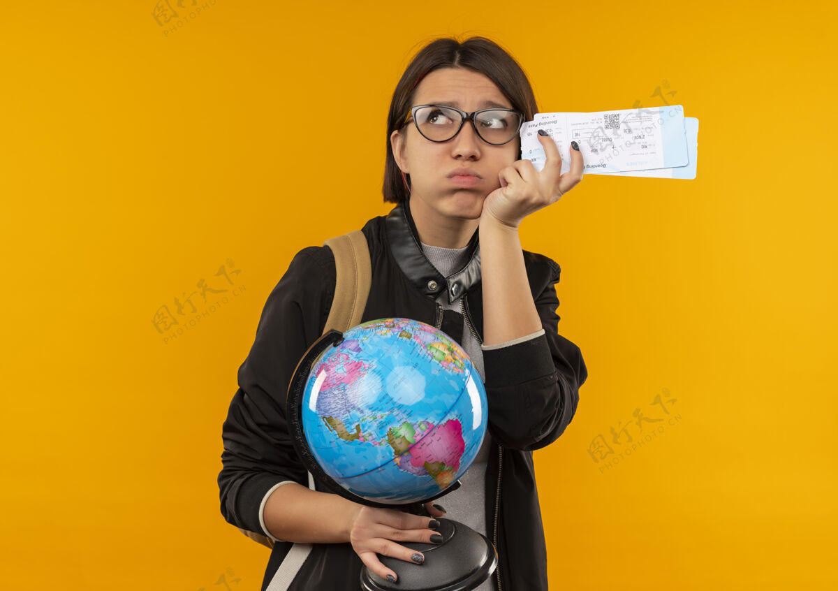 地球仪未出租的年轻学生女孩戴着眼镜 背着包拿着飞机票和地球仪 一边看着一边看着一边隔离在橙色背景上的复印空间背部飞机持有