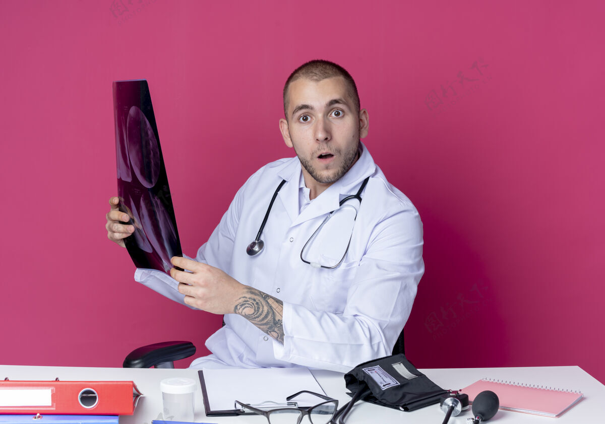 X光令人印象深刻的年轻男医生穿着医用长袍和听诊器坐在办公桌旁 拿着工作工具 拿着x光照片 看着前面的粉红色隔离拿着工具医疗