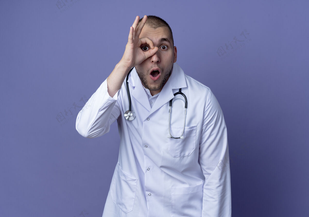 医生印象深刻的年轻男医生穿着医用长袍和听诊器做着孤立的紫色上看手势医疗男性听诊器