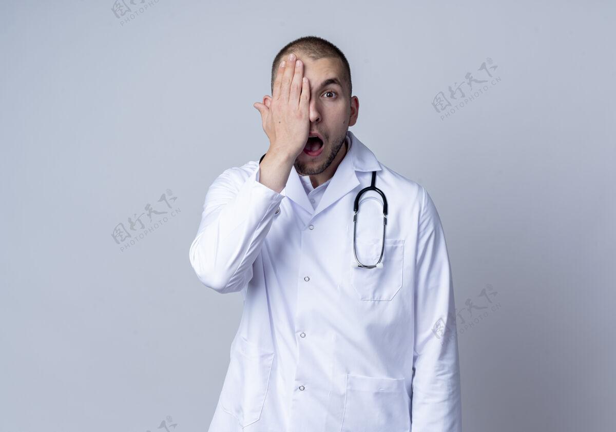 听诊器令人印象深刻的年轻男医生穿着医用长袍 脖子上戴着听诊器 用手遮住半张脸半戴印象