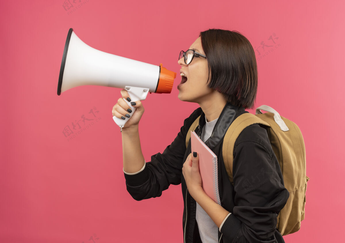 持有戴着眼镜背着书包的年轻女学生站在侧视图里拿着便笺本说话被隔离在粉红色的讲话者包粉红色背部