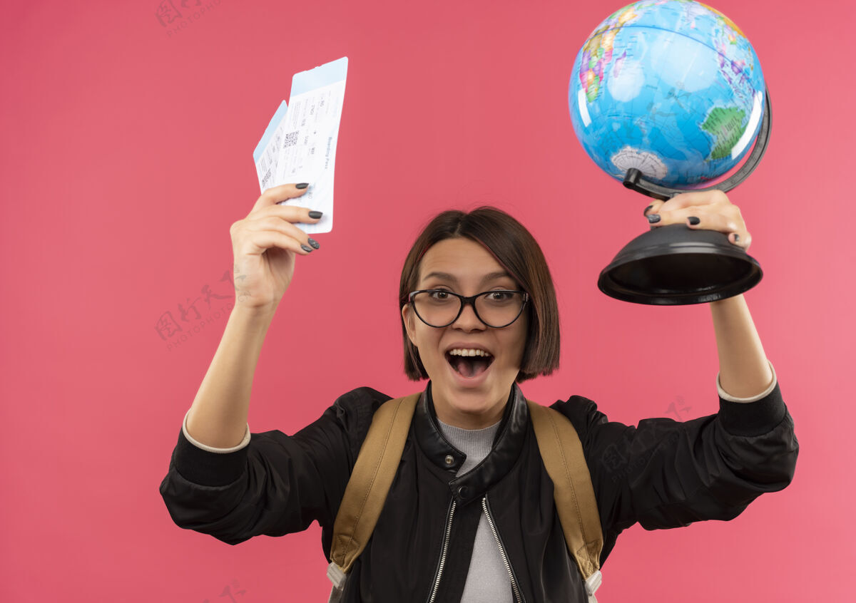 喜悦快乐的年轻学生女孩戴着眼镜 背着提包举起飞机票和地球仪孤立在粉红色回来地球仪眼镜