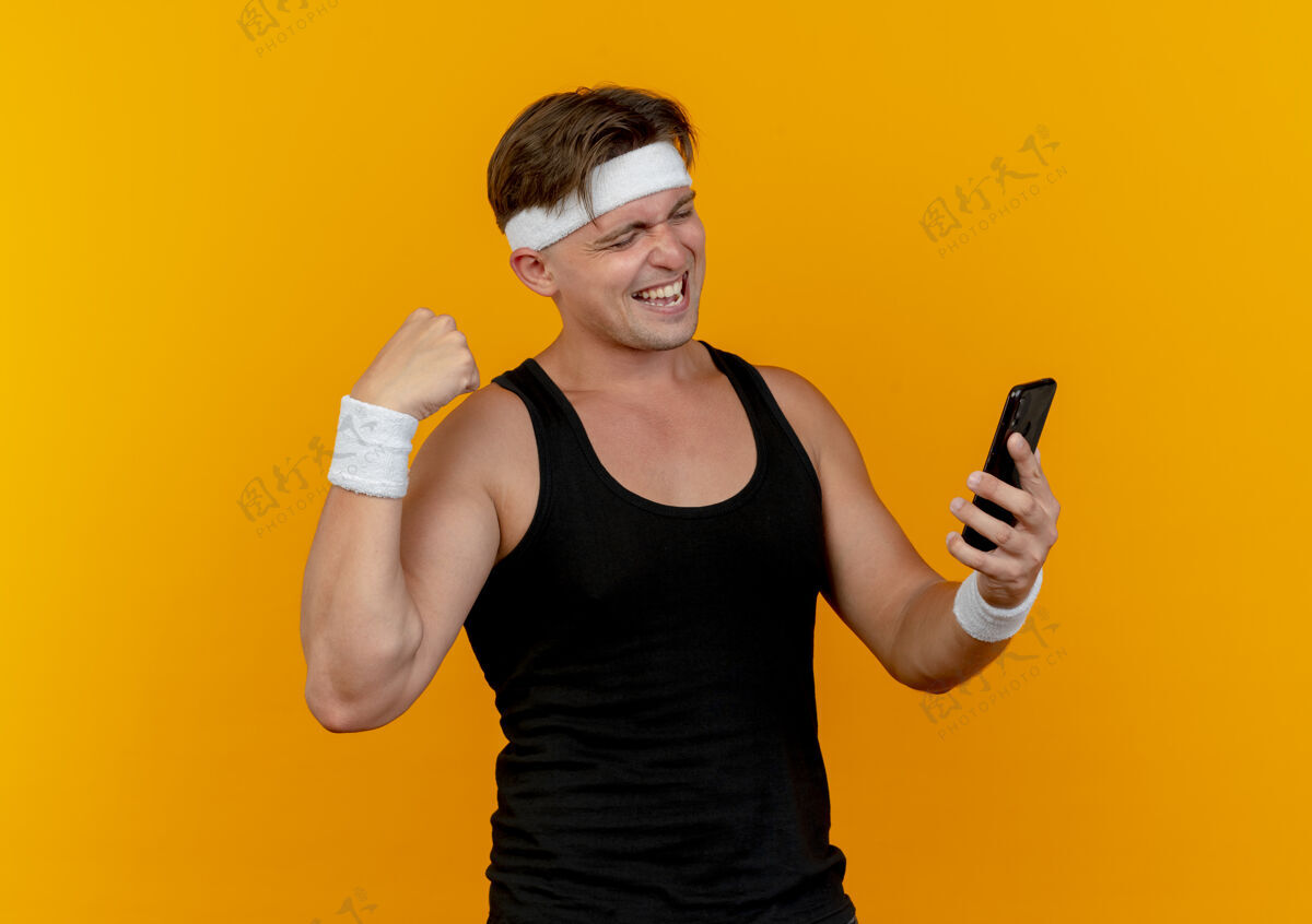 手势快乐的年轻帅气的运动型男人戴着头带和腕带拿着手机 看着手机 在橙色上做着强烈的孤立的手势腕带男士手持