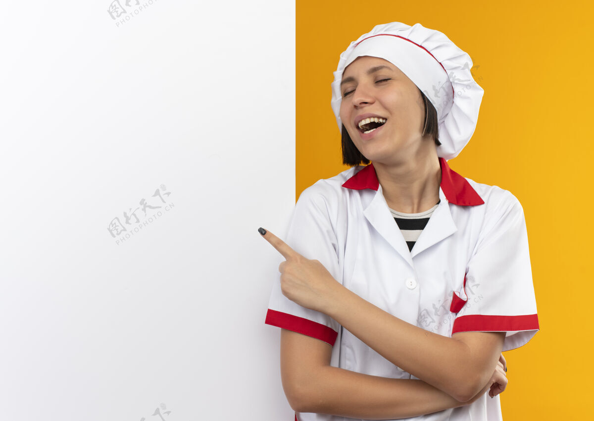 站着身着厨师制服的年轻快乐的女厨师站在白色的墙前 闭着眼睛指着墙 眼睛被橘色隔离制服厨师女性