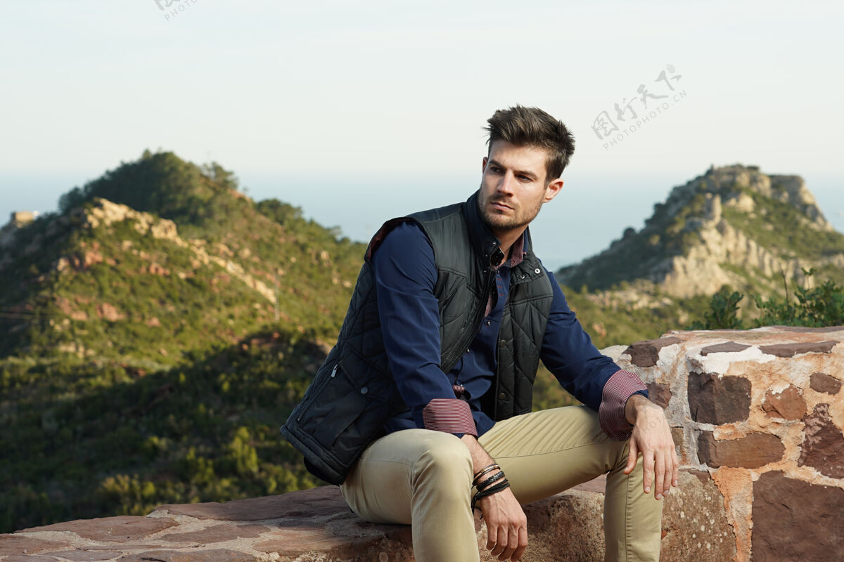 表面穿着蓝色衬衫和黑色夹克的年轻西班牙裔男模在石墙旁摆姿势石头天气绅士