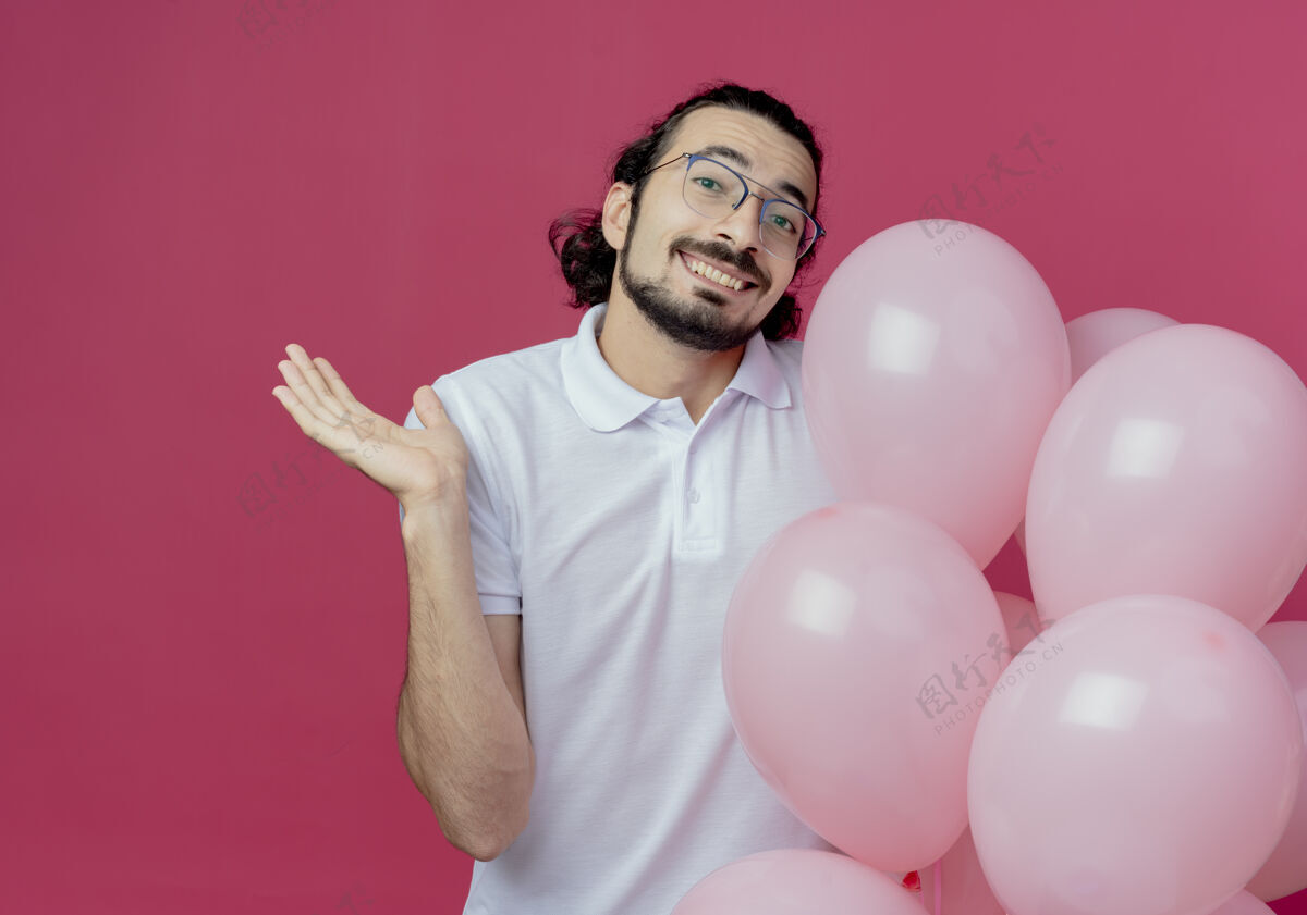 帅气笑容可掬的帅哥戴着眼镜拿着气球 用手指着隔离在粉色背景上的复制空间微笑穿拿着
