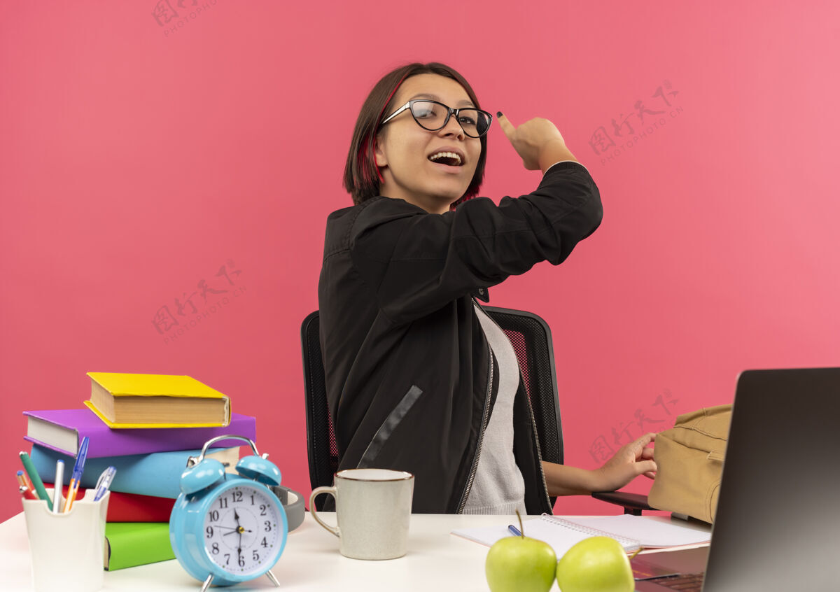 印象给人印象深刻的年轻女学生戴着眼镜坐在书桌旁做作业 指着后面孤立的粉红色眼镜戴着粉色
