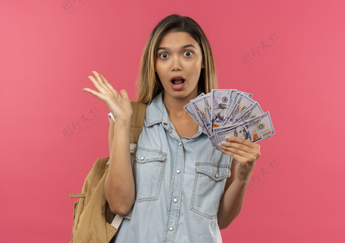 秀印象深刻的年轻漂亮的学生女孩穿着背袋拿着钱 并显示空的手上孤立的粉红色抱空背
