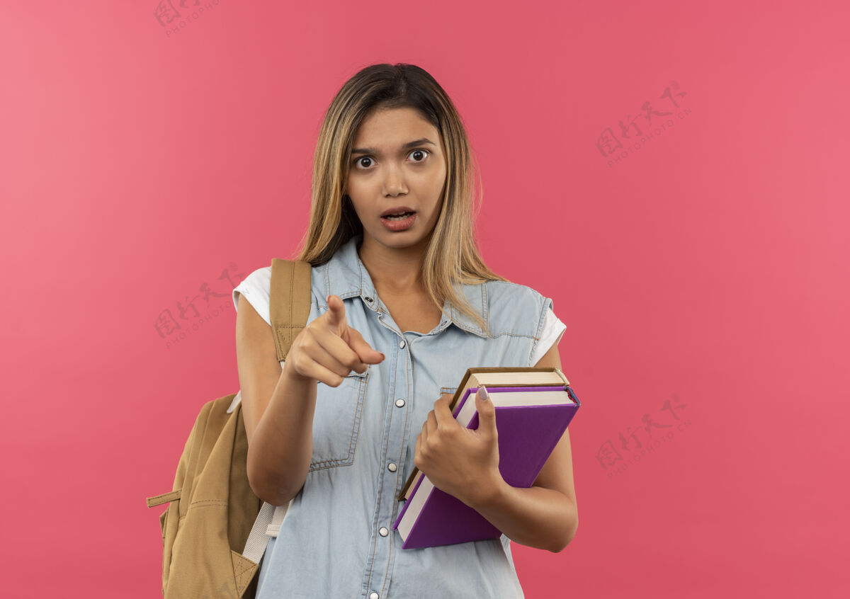 学生印象深刻的年轻漂亮的学生女孩穿着背包拿着书 指着前面孤立的粉红色年轻粉色指着