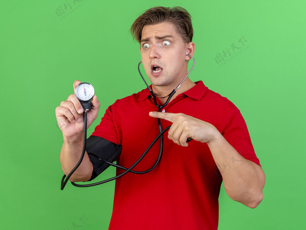 站着印象深刻的年轻英俊的金发病男子戴着听诊器测量压力给自己拿着看 指着隔离在绿色墙上的血压计他自己人市民