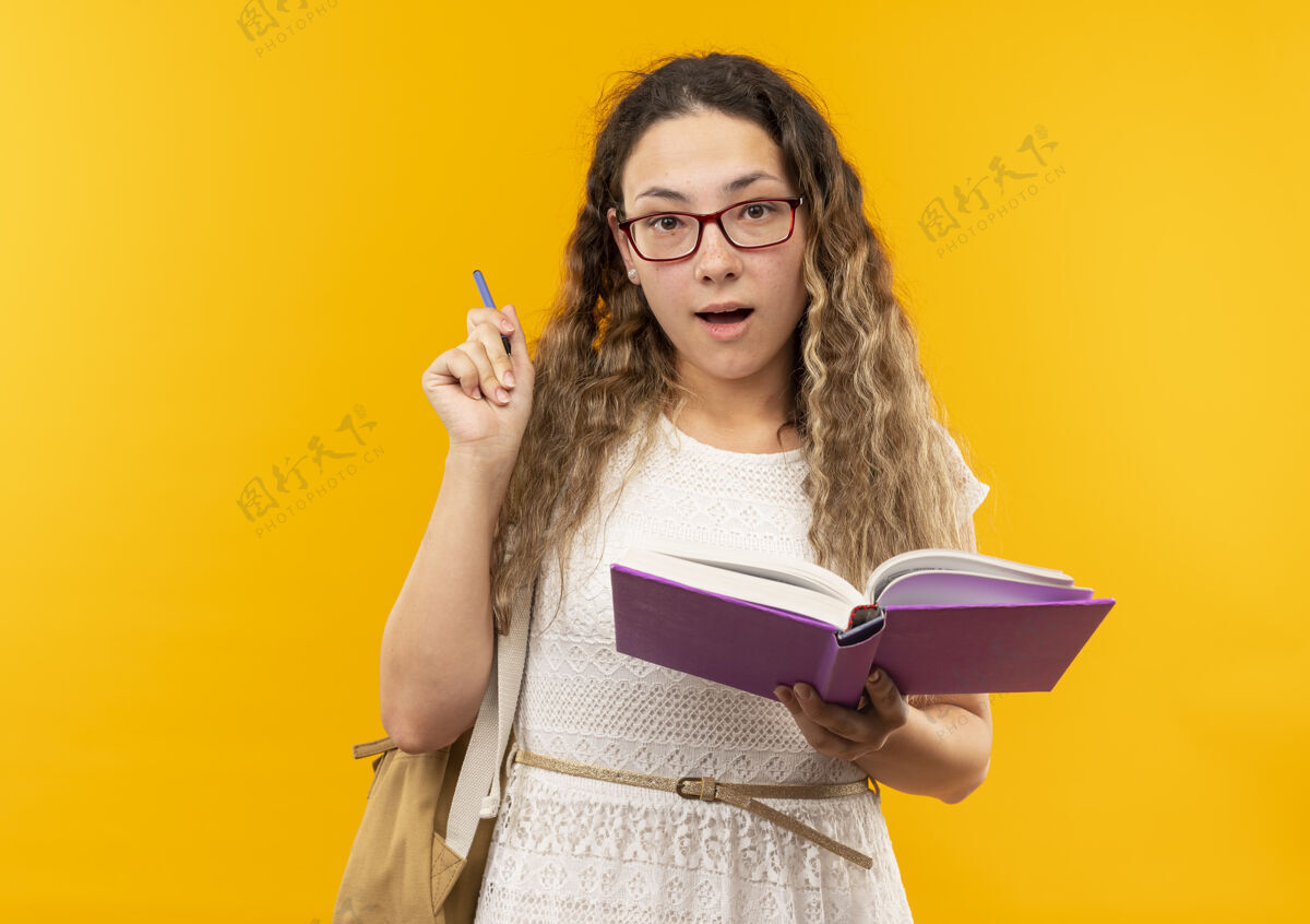 包印象深刻的年轻漂亮的女学生戴着眼镜 背着书包拿着笔和书孤立在黄色的眼镜黄色印象