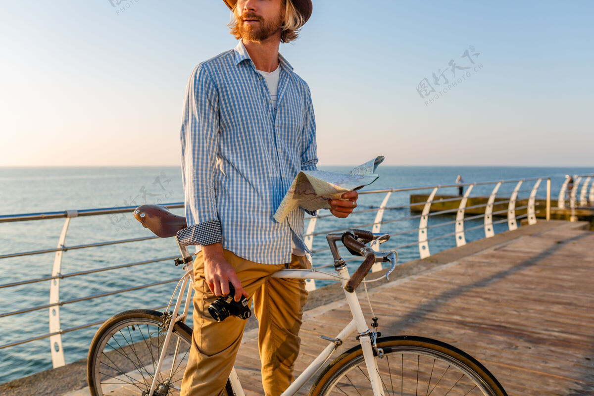 日落年轻帅哥暑假骑着自行车在海边旅游 日落时分在海边 波西米亚时髦的装扮 拿着地图观光拍照 穿着衬衫和帽子自行车户外骑