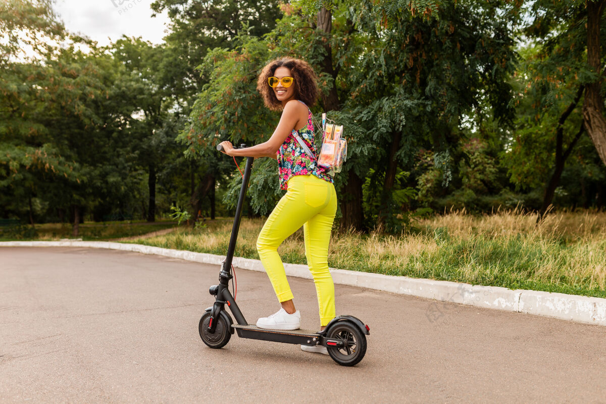 太阳镜年轻时尚的黑人女子在公园里玩得很开心 骑着电动脚踏车 夏日时尚 五颜六色的时髦装扮 背着背包 穿着黄色裤子和太阳镜电力人车辆