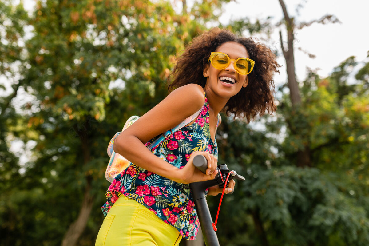 道路年轻时尚的黑人女子在公园里玩得很开心骑着电动脚踏车在夏天的时尚风格 五颜六色的时髦服装 穿着背包和黄色太阳镜女孩人车辆