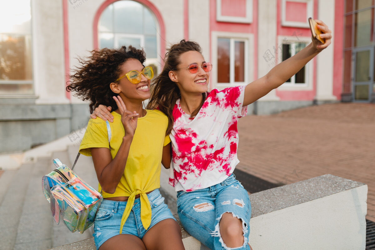 非裔美国人五颜六色的照片 快乐的年轻女孩朋友们微笑着坐在街上用手机自拍 女人们在一起玩得很开心欢呼眼镜兴奋