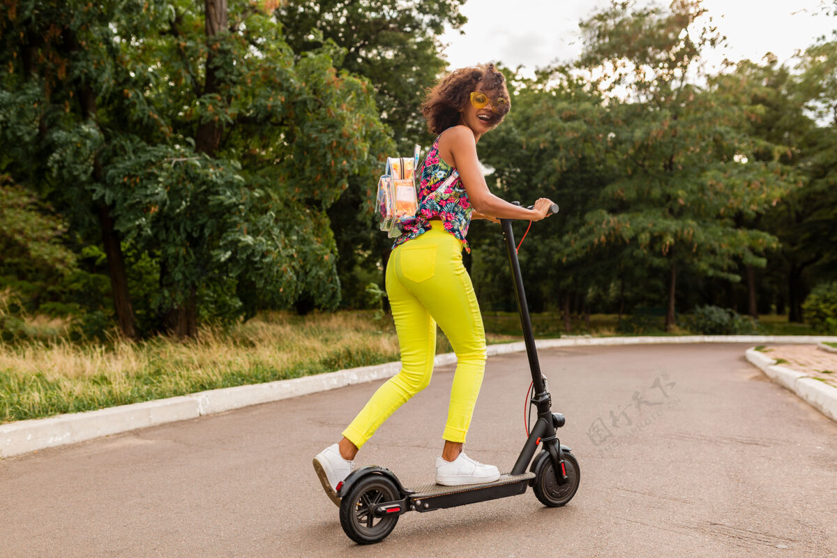 环保年轻时尚的黑人女子在公园里玩得很开心骑着电动脚踏车在夏天的时尚风格 五颜六色的时髦服装 穿着背包和黄色太阳镜微笑兴奋女士
