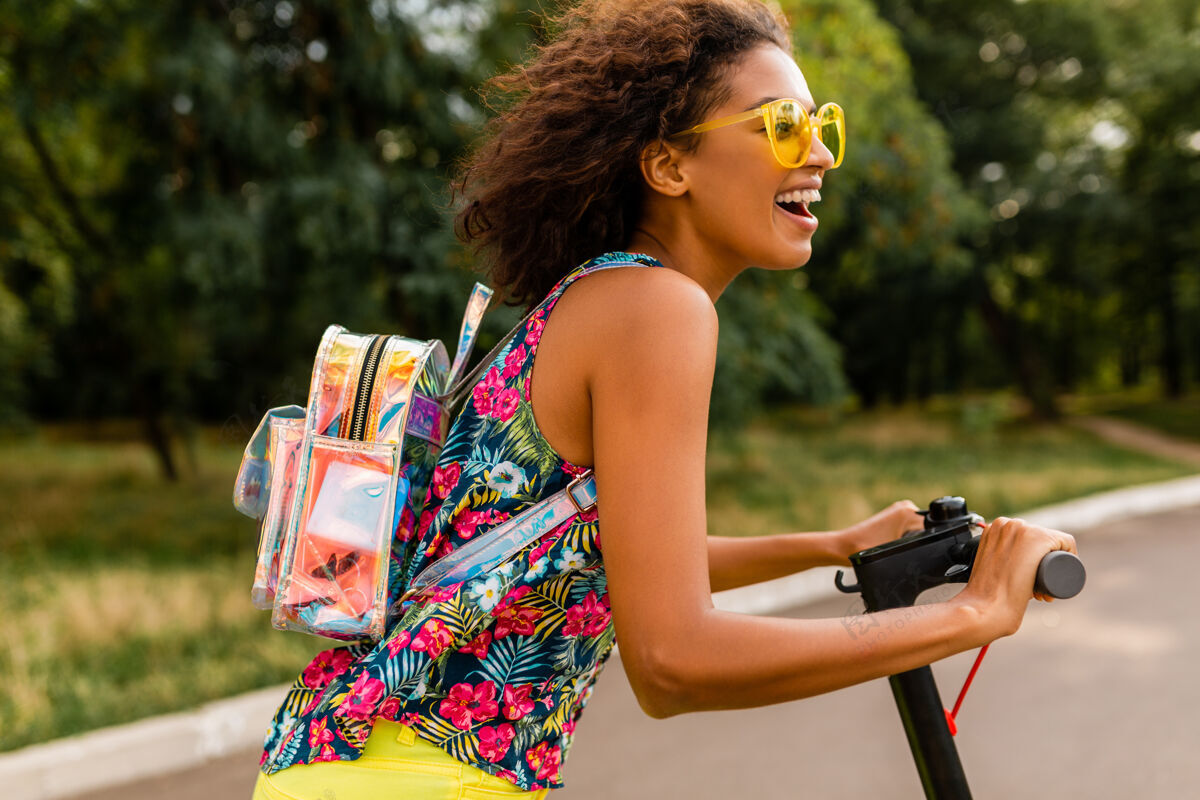 乘坐年轻时尚的黑人女子在公园里玩得很开心骑着电动脚踏车在夏天的时尚风格 五颜六色的时髦服装 穿着背包和黄色太阳镜女人多彩兴奋