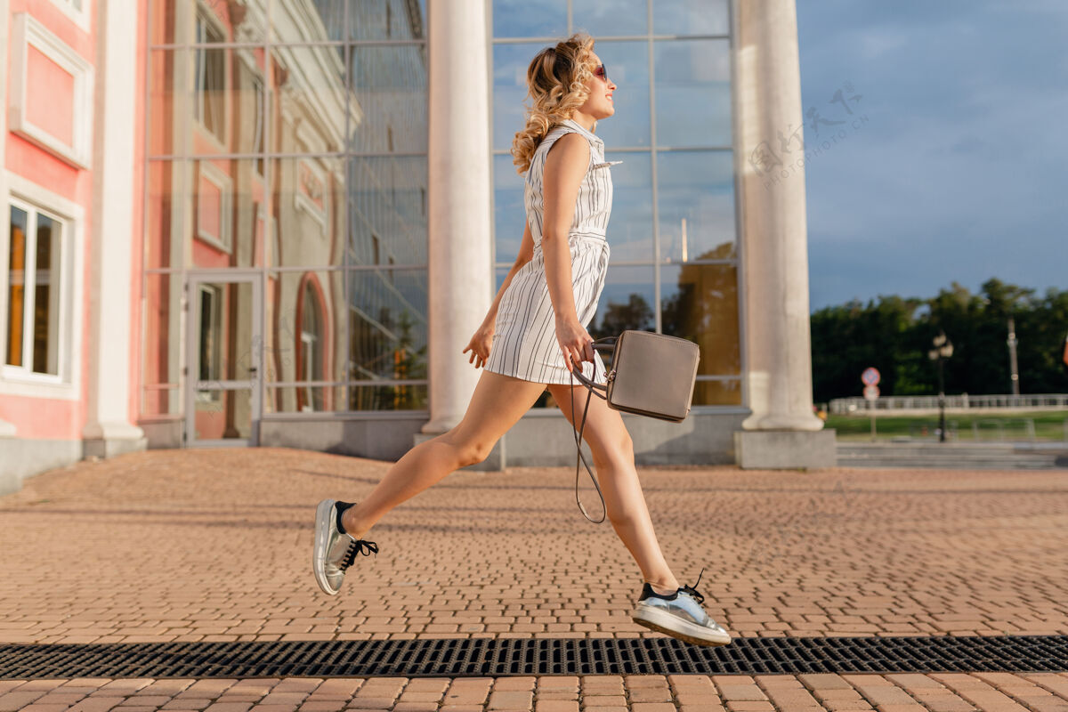 女性年轻迷人的时尚女子穿着运动鞋在城市街道上奔跑跳跃有趣夏天时尚风格的白色连衣裙戴着太阳镜和手提包优雅连衣裙姿势