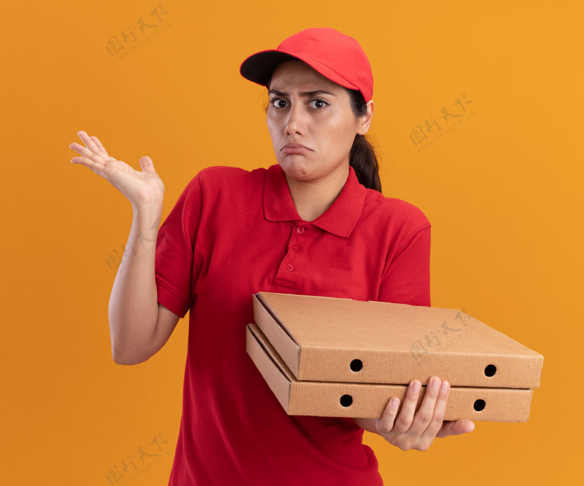戴着困惑的年轻送货女孩穿着制服 戴着帽子 手里拿着披萨盒 手被隔离在橙色的墙上摊开送货帽子