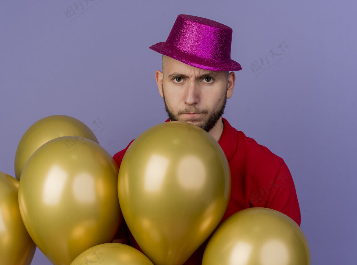 派对年轻帅气的斯拉夫党人戴着党帽站在气球后面看着紫色背景上孤立的摄像机小伙子紫色讨厌的