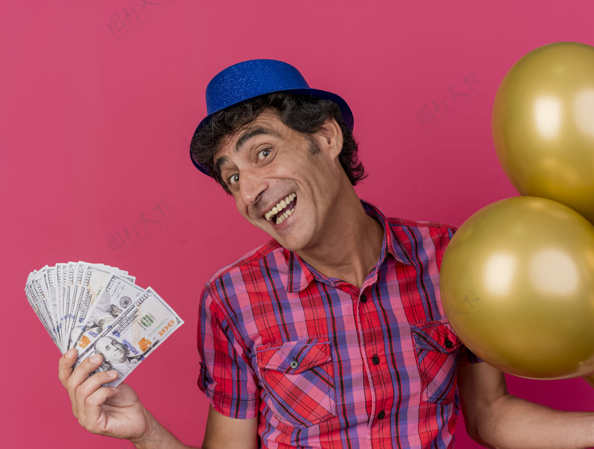 举行快乐的中年白种人党男子戴着党的帽子拿着气球和钱看着摄像机隔离在深红色的背景男人帽子中年