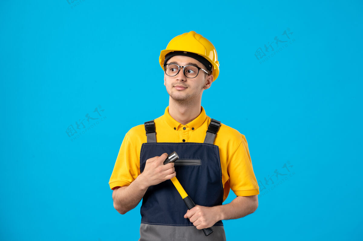 工作蓝色墙壁上穿着制服手持锤子的男建筑工人的正视图帽子制服前面