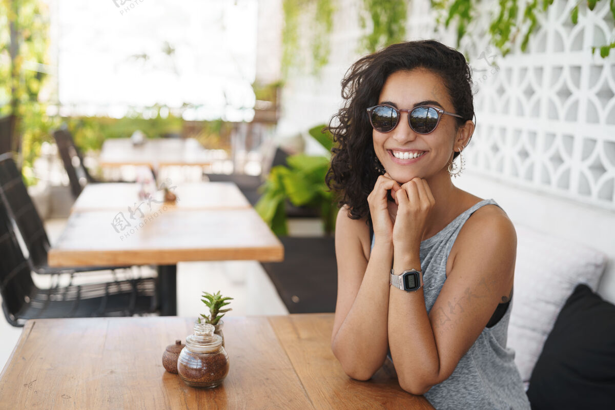 独自戴着墨镜的快乐女人坐在咖啡馆的夏日露台上杯子周末庭院