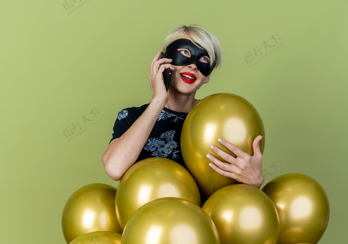 抢夺令人印象深刻的年轻金发派对女孩戴着化装面具站在气球后面抓住其中一个看着一边讲电话孤立在橄榄绿的背景化装舞会派对面具