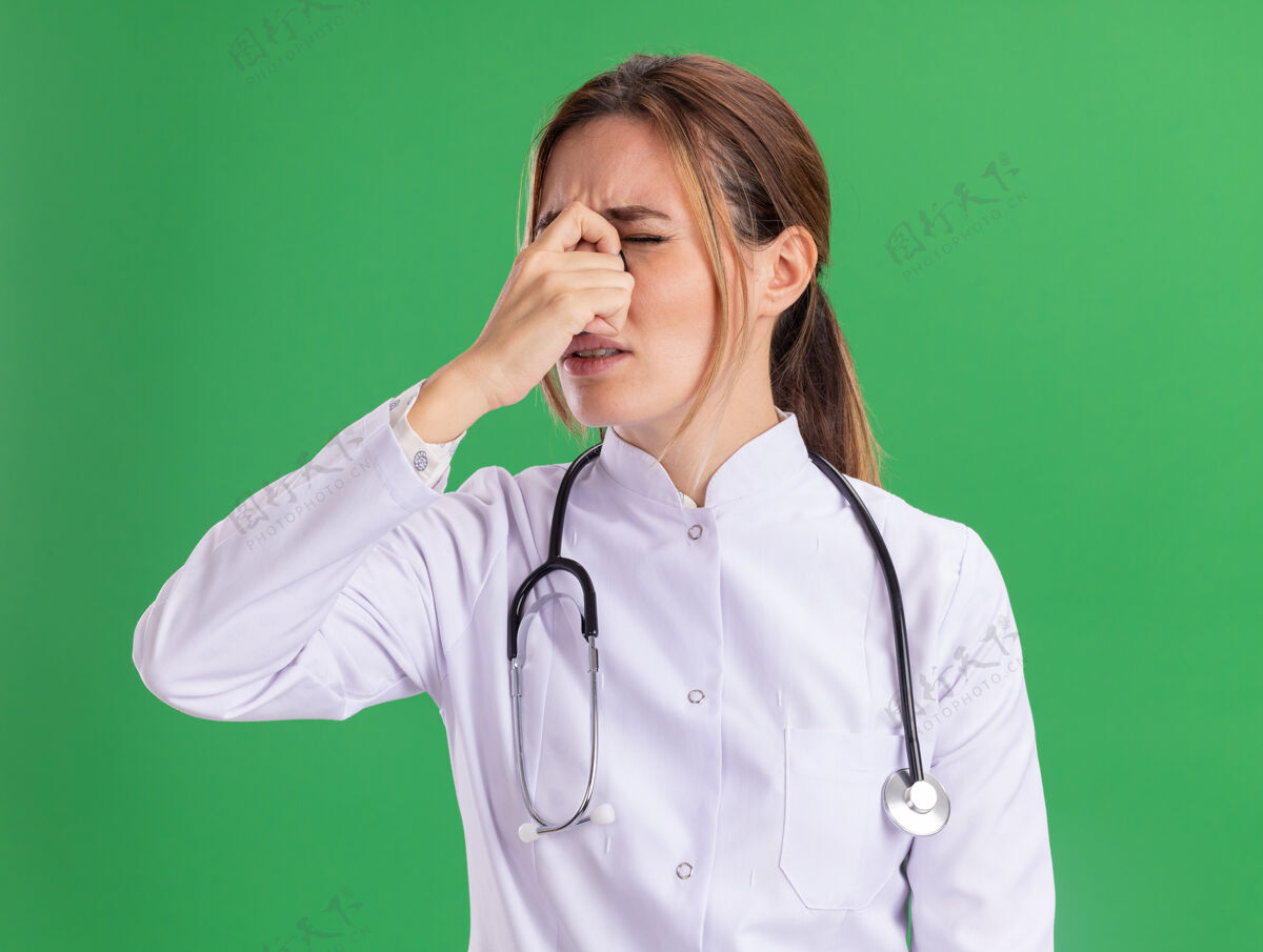 医生厌倦了闭着眼睛的年轻女医生穿着医用长袍 用听诊器抓住了隔离在绿墙上的鼻子疲倦女性抓