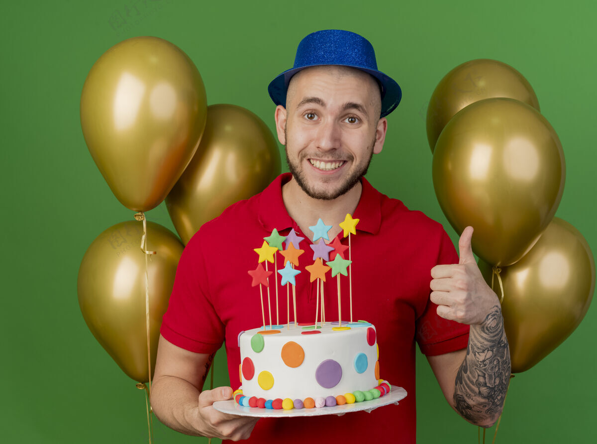 斯拉夫面带微笑的年轻帅哥斯拉夫人戴着派对帽站在气球前拿着生日蛋糕竖起大拇指看着隔离在绿色背景上的相机抱着绿色站着