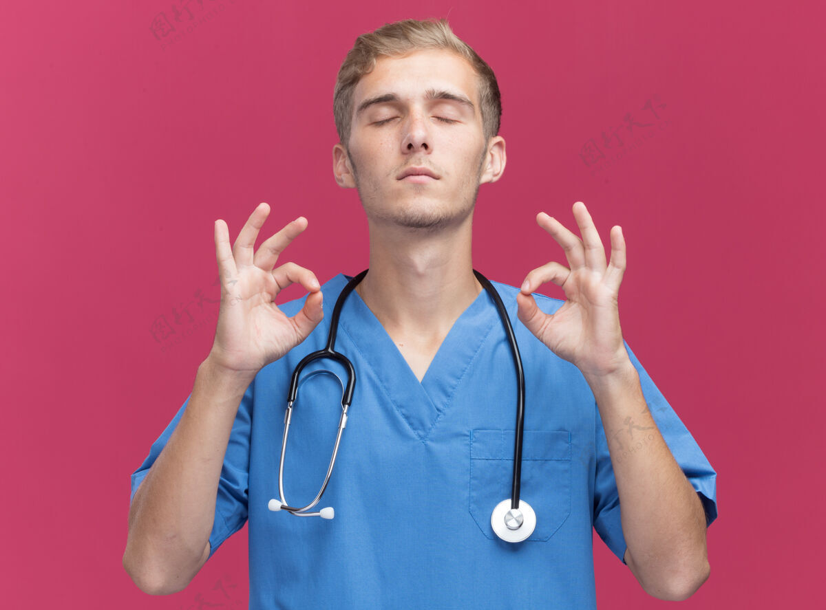 关闭年轻的男医生闭着眼睛 穿着医生制服 带听诊器 在粉红色的墙上显示冥想的姿势年轻手势医生