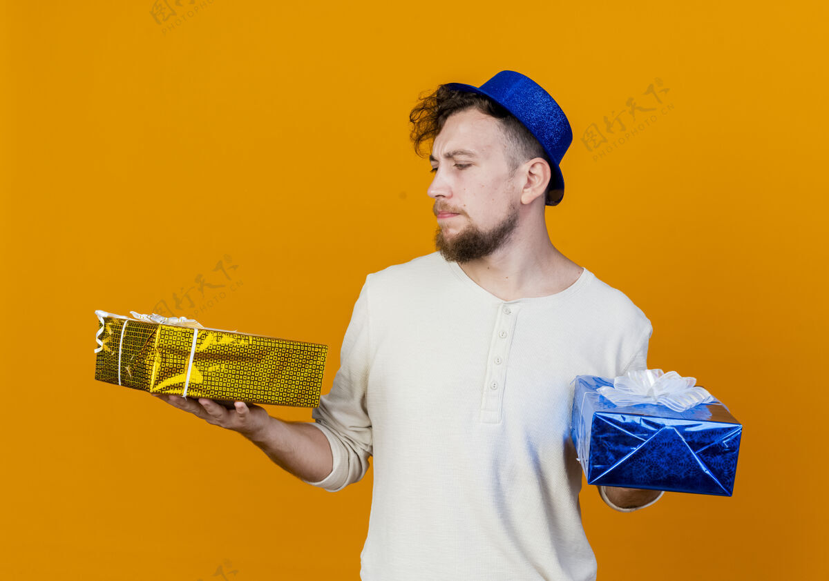 斯拉夫人一个年轻英俊的斯拉夫人 戴着派对帽 拿着礼物盒 看着橙色背景的礼物盒抱着年轻人派对