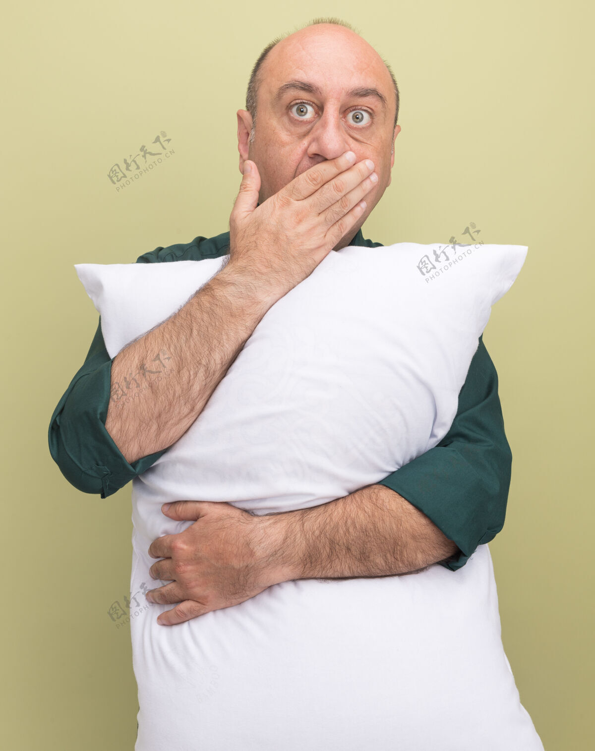 橄榄惊讶的中年男子穿着绿色t恤 抱着枕头捂着嘴 手被隔离在橄榄绿的墙上T恤中年枕头