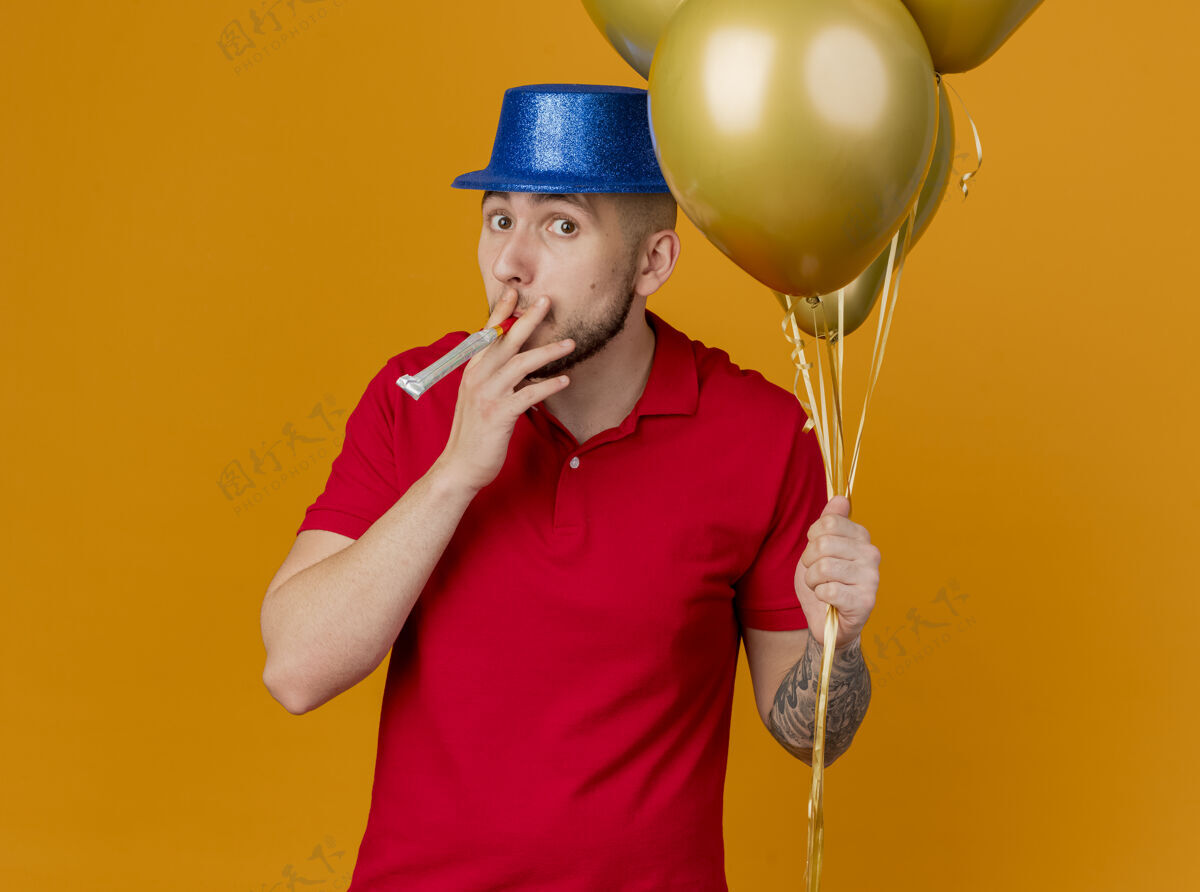 帽子令人印象深刻的年轻英俊的斯拉夫党人戴着党的帽子拿着气球看着相机吹党鼓风机孤立的橙色背景吹气球印象