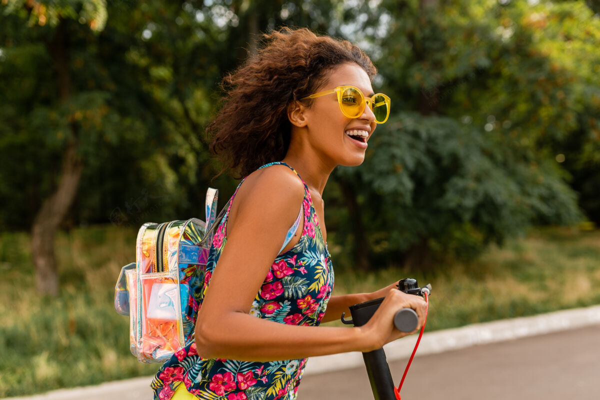埃斯库特年轻时尚的黑人女子在公园里玩得很开心骑着电动脚踏车在夏天的时尚风格 五颜六色的时髦服装 穿着背包和黄色太阳镜电力女性街道