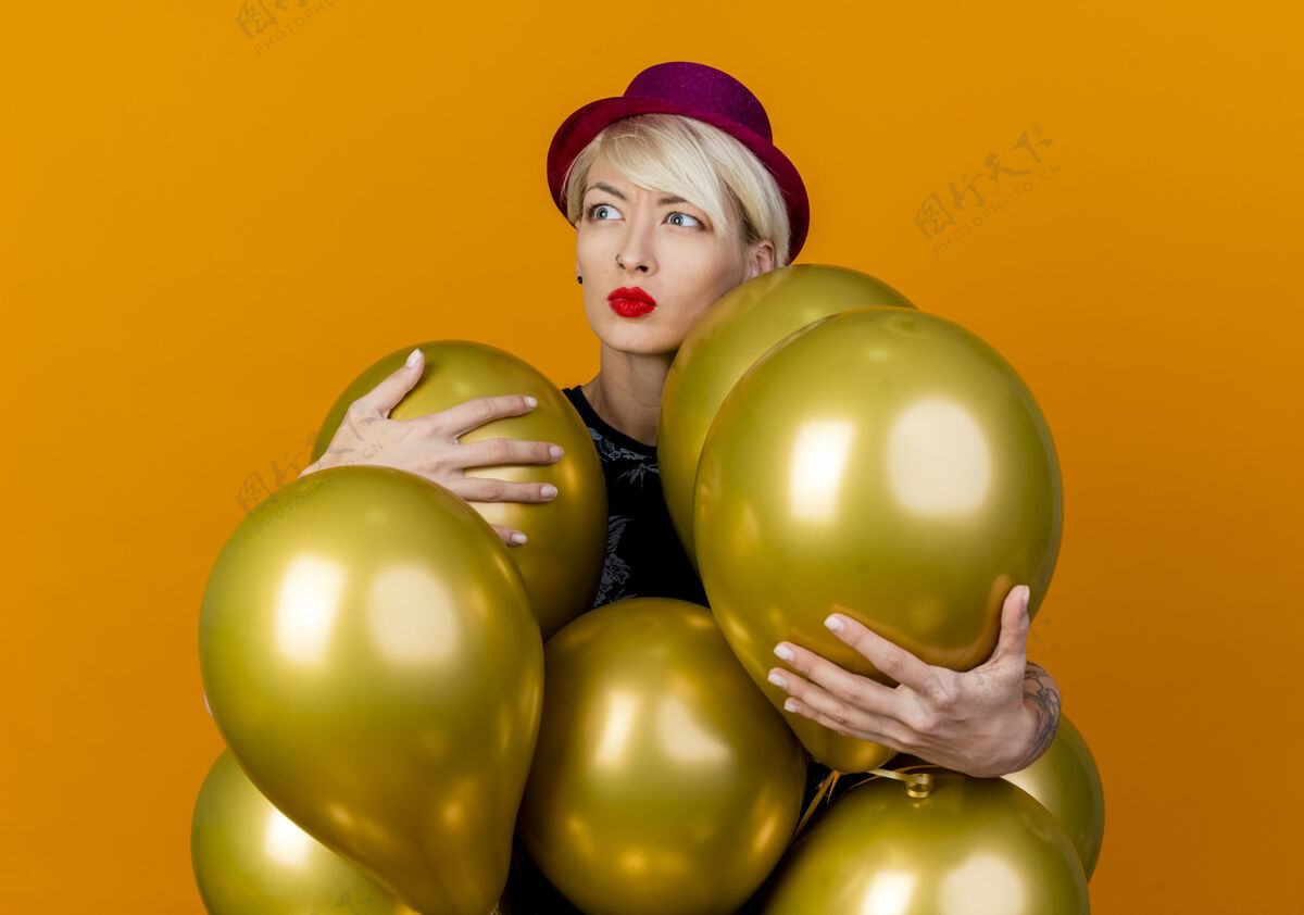 气球皱眉的年轻金发派对女郎戴着派对帽站在气球后面抓着气球看着橙色背景下孤立的一面帽子女孩年轻