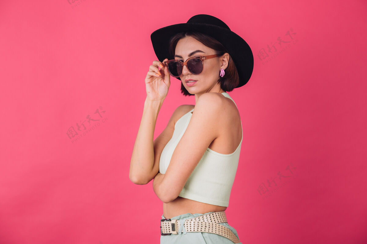 帽子戴着帽子和太阳镜的漂亮时髦女人在粉红色的墙上摆姿势唇膏嘴唇成人