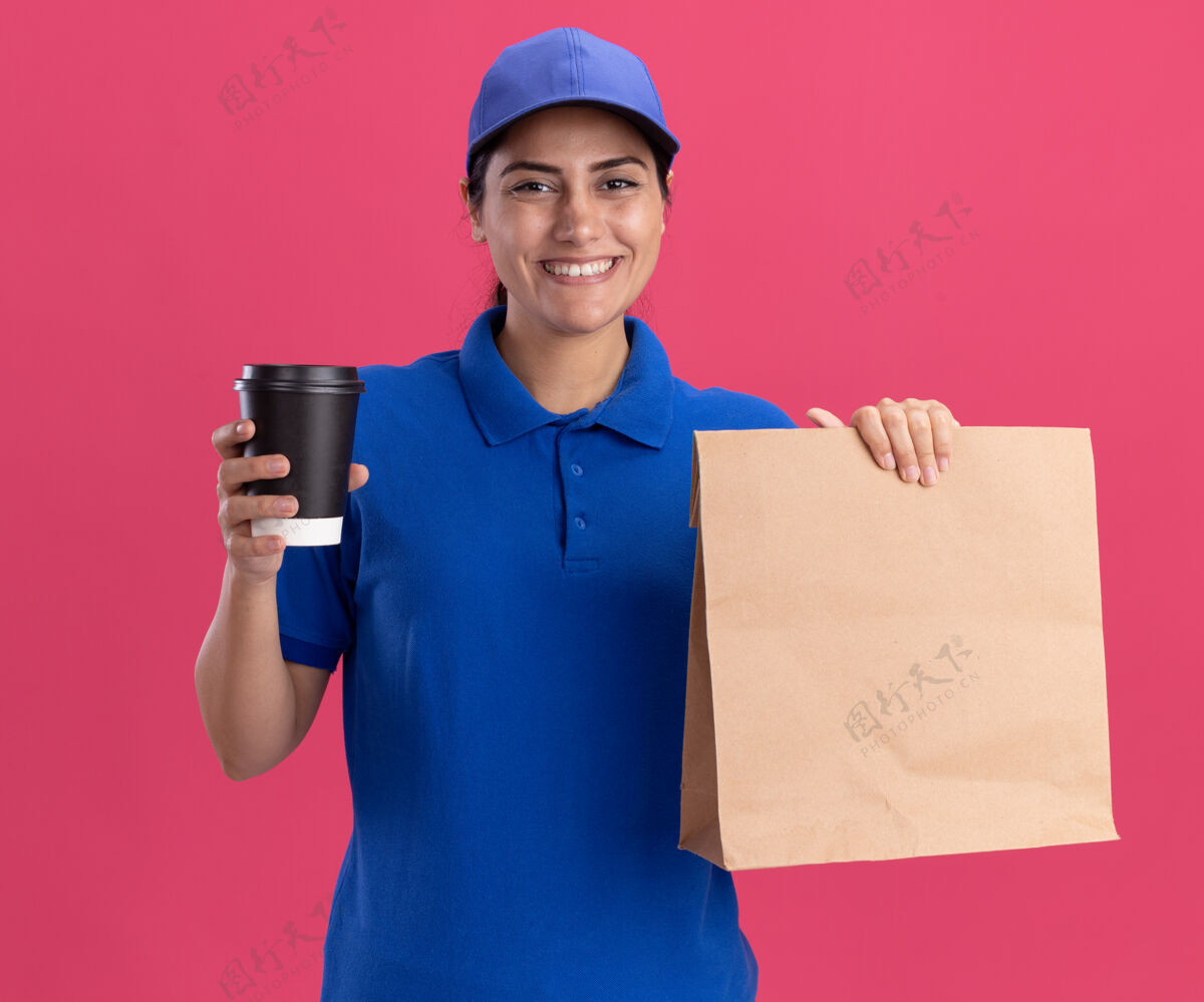 食物带着微笑的年轻送货女孩 穿着制服 戴着帽子 手里拿着一个纸食品包 粉红色的墙上隔着一杯咖啡纸咖啡穿着