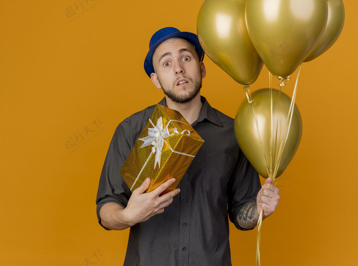 年轻令人印象深刻的年轻英俊的斯拉夫党的家伙戴着党的帽子拿着气球和礼品包看着相机孤立的橙色背景与复制空间帽子举行包