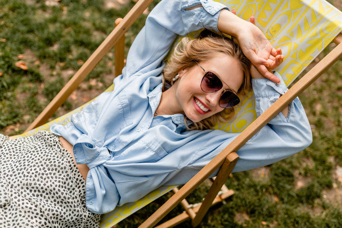 快乐迷人的金发微笑的女人坐在躺椅上穿着夏天的衣服头发包美丽
