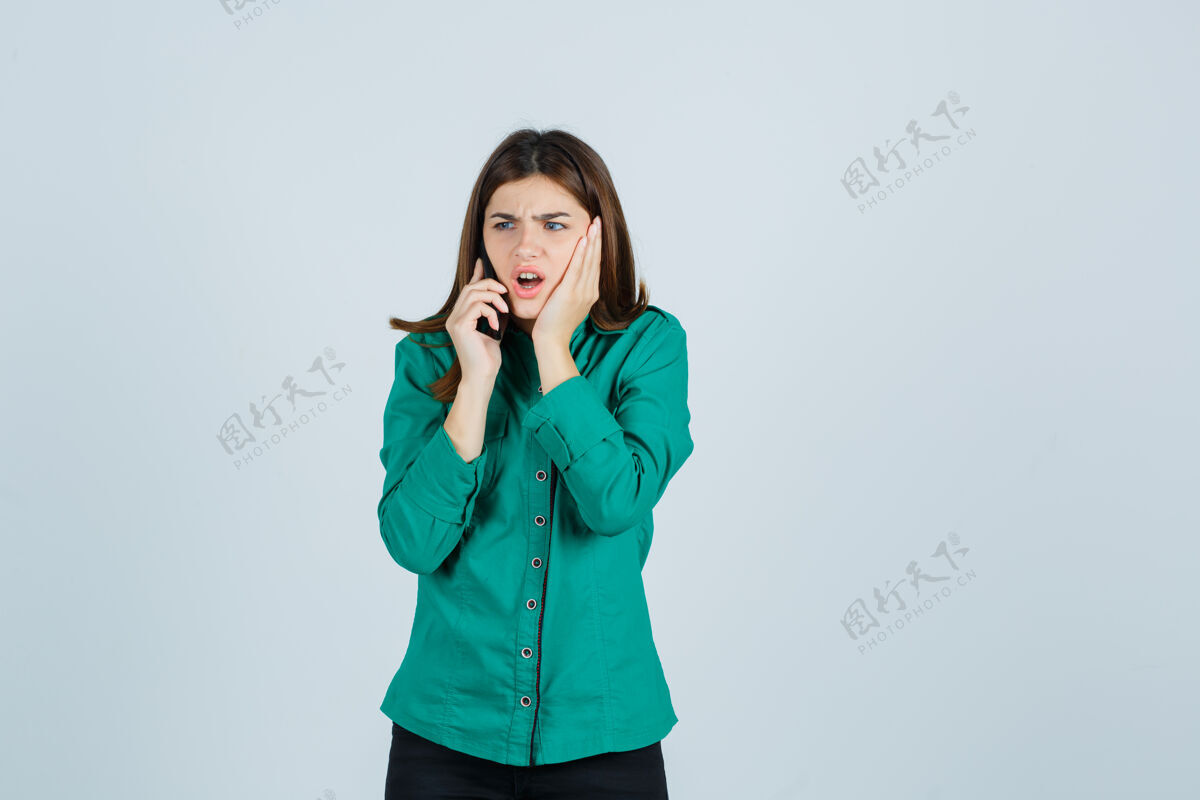 裤子穿着绿色上衣 黑色裤子的年轻女孩在和电话交谈 手放在脸颊上 看上去很震惊开放年轻电话