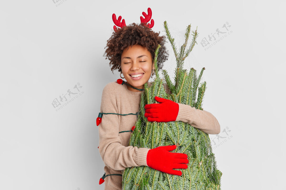 购买笑容可掬的美国黑人妇女戴着驯鹿的角和手套怀着爱拥抱着绿杉树快乐地在家里庆祝新年从圣诞街市场回来用花环包着拥抱满意季节