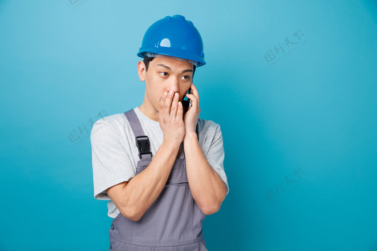 制服印象深刻的年轻建筑工人戴着安全帽和制服耳语在电话边看建筑复制年轻