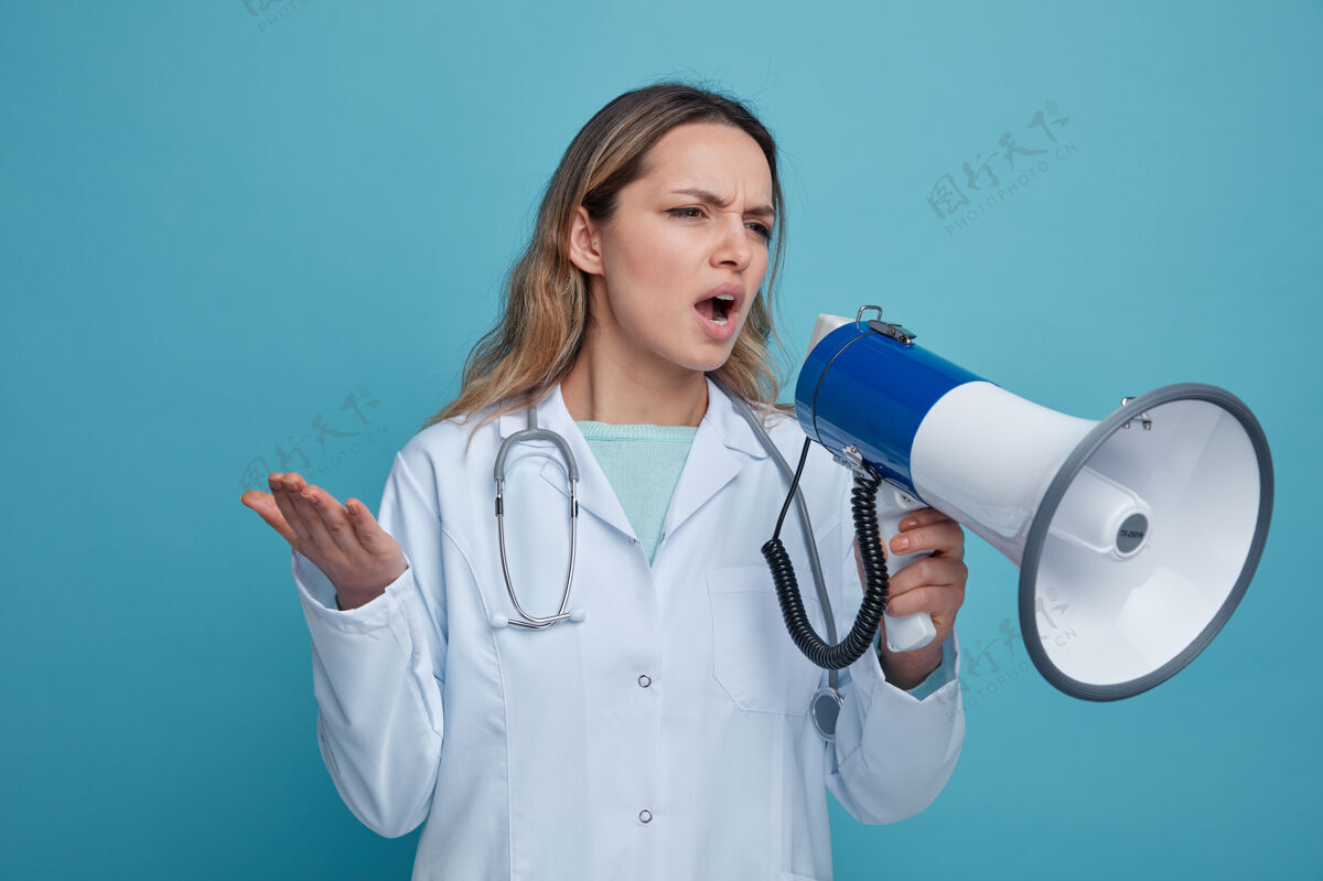显示恼怒的年轻女医生穿着医用长袍 脖子上戴着听诊器 一边看一边说话 说话的人空手而出手穿壁板